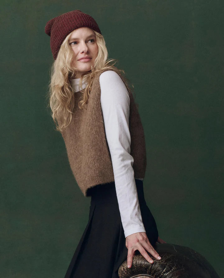 Woman wearing a brown knitted vest, white shirt, and red beanie against a green background, side