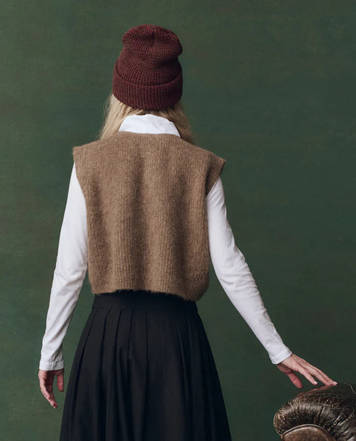 Person wearing a brown knitted vest, white shirt, and dark skirt against a green background, back