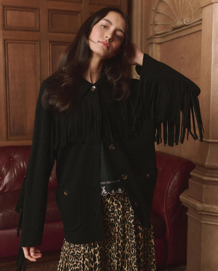 A woman wearing a black fringed jacket with buttons and a leopard-print skirt, posing in front of a wooden-paneled wall and a red leather sofa.