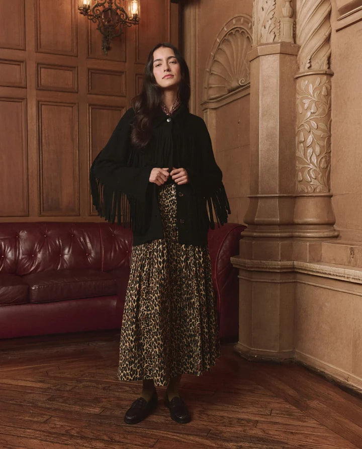 A woman stands in an ornate indoor room with wood-paneled walls, a red leather sofa, and carved columns, wearing a black fringed cardigan over a leopard-print midi skirt with dark loafers.