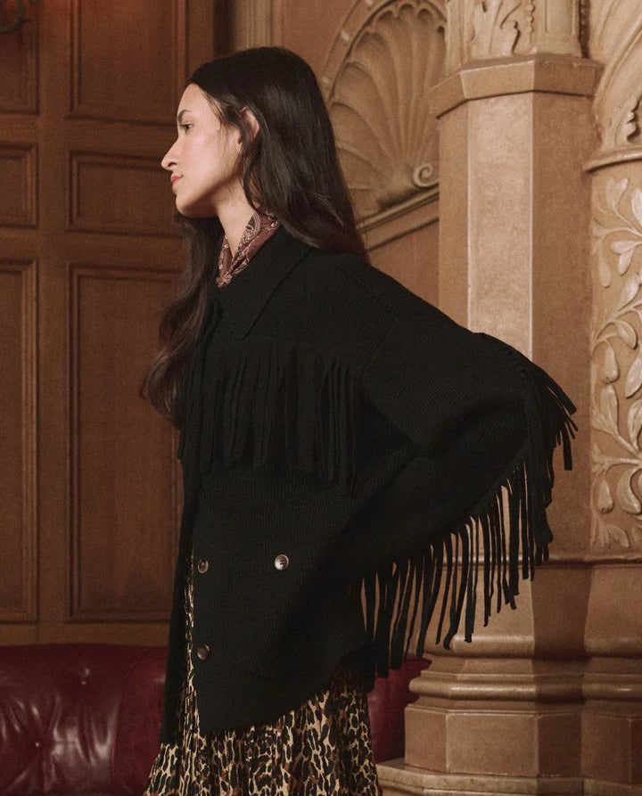 A woman in profile wearing a black fringed cardigan over a leopard-print dress, standing in an ornate indoor setting with wood paneling and carved columns.