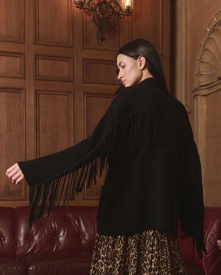 A woman wearing a black fringed jacket, shown from the back in an indoor setting with wood-paneled walls and a red leather sofa.