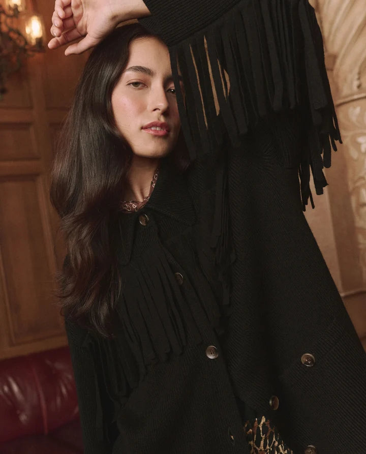 A woman wearing a black fringed coat with buttons, posed in a warm, elegant indoor setting with wood paneling and a red leather seat visible in the background.