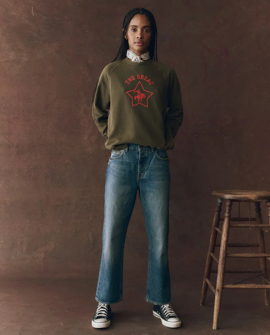 The college sweatshirt rodeo star army green on a model wearing jeans with a brown background, front view