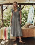 Woman in a checkered dress standing in a rustic outdoor setting with trees and a wooden structure.