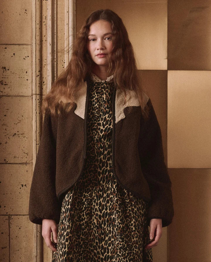 A woman wearing a dark brown shearling jacket with beige collar panels over a leopard-print dress, standing against a warm beige tiled wall.