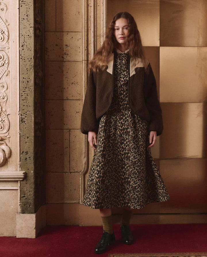 A model stands in an ornate interior wearing a brown suede jacket with a beige shearling collar over a leopard-print midi dress, paired with olive socks and black shoes.