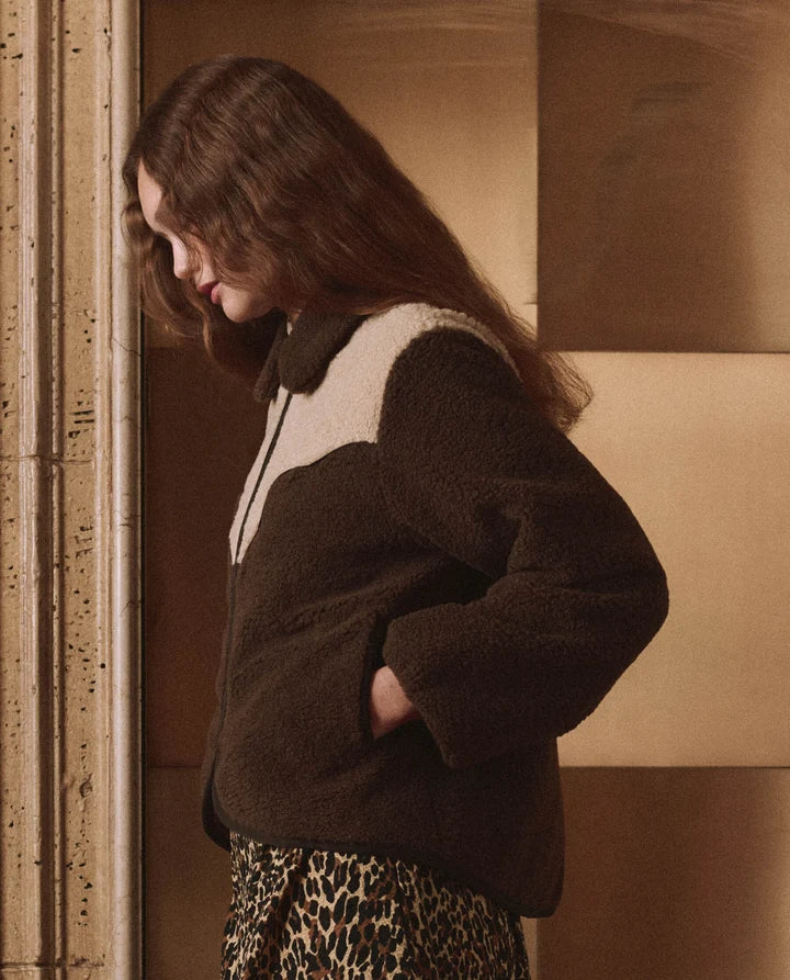 A woman wearing a dark brown sherpa fleece jacket with cream color blocking and a leopard-print skirt, standing in a warm-toned interior setting.