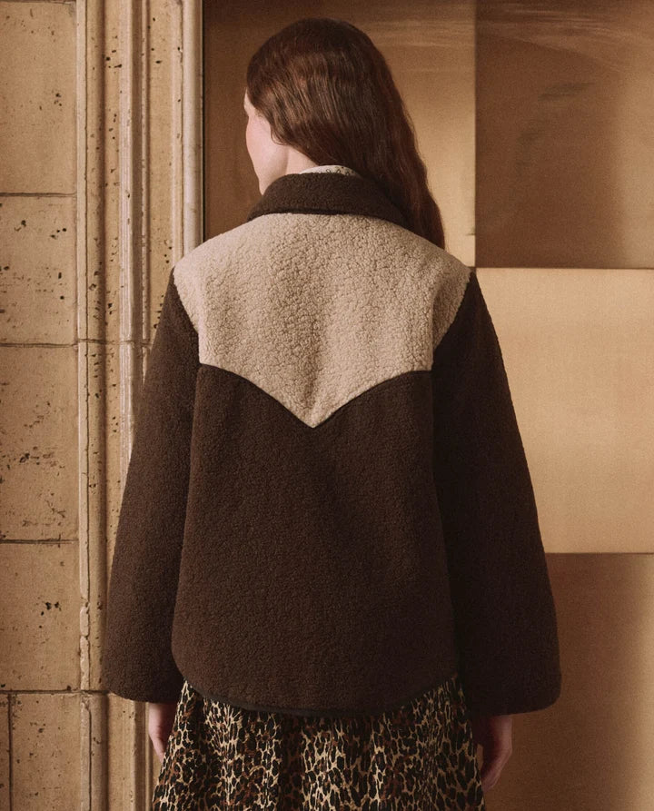 A model viewed from behind wearing a dark brown teddy fleece jacket with a contrasting beige shearling-style yoke panel, paired with a leopard-print skirt, set against a warm-toned architectural background.