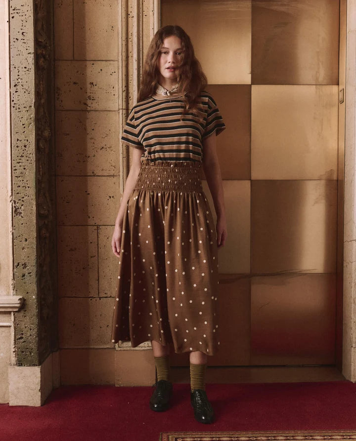 A woman stands indoors wearing a brown and black striped short-sleeve top with a brown polka-dot midi skirt, olive green socks, and black shoes, posed in front of a warm-toned tiled wall and red carpet.