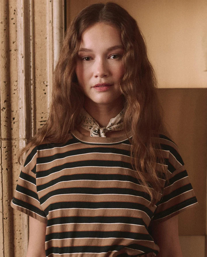 A woman with long wavy light brown hair wearing a brown and black striped short-sleeve top with a patterned neck scarf, standing in a warm-toned indoor setting.