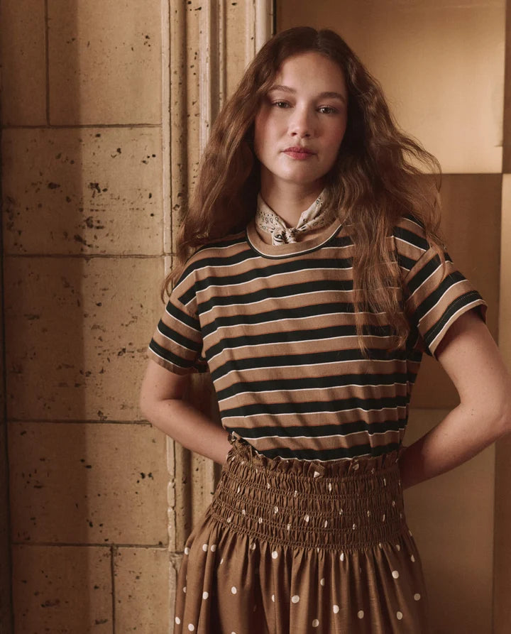 A woman with long wavy light brown hair stands indoors wearing a beige and black horizontally striped short-sleeve top with a patterned neck scarf, paired with a brown smocked skirt with white polka dots.
