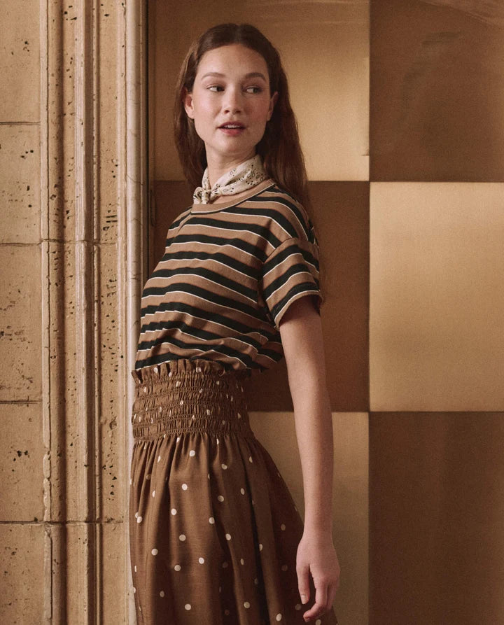 A woman modeling a retro-style outfit consisting of a brown and black striped short-sleeve top with a cream neck scarf, paired with a brown smocked skirt with white polka dots, set against a warm checkered wall.