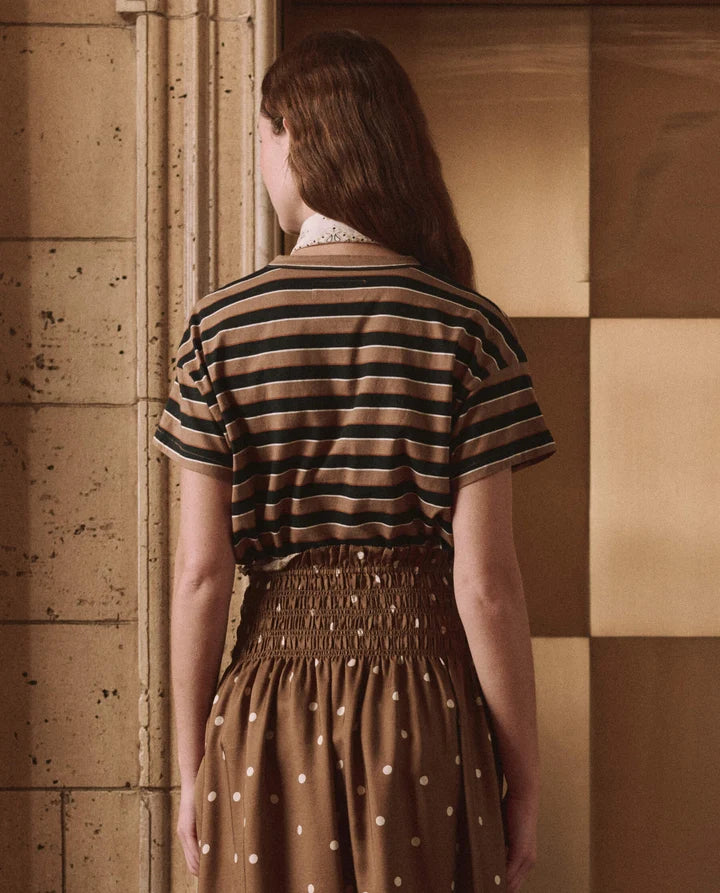 A woman viewed from behind wearing a brown and black striped short-sleeve top and a brown smocked skirt with white polka dots, standing against a warm-toned paneled wall.