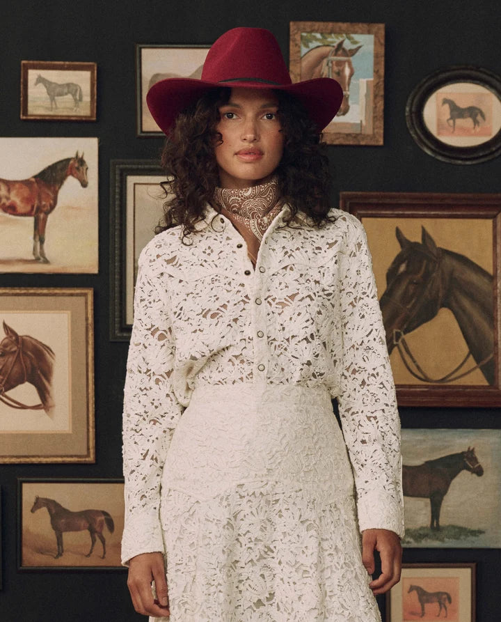 A woman stands indoors wearing a white lace long-sleeve shirt and matching skirt with a dark red wide-brim hat. The background is a dark wall covered with framed horse paintings.