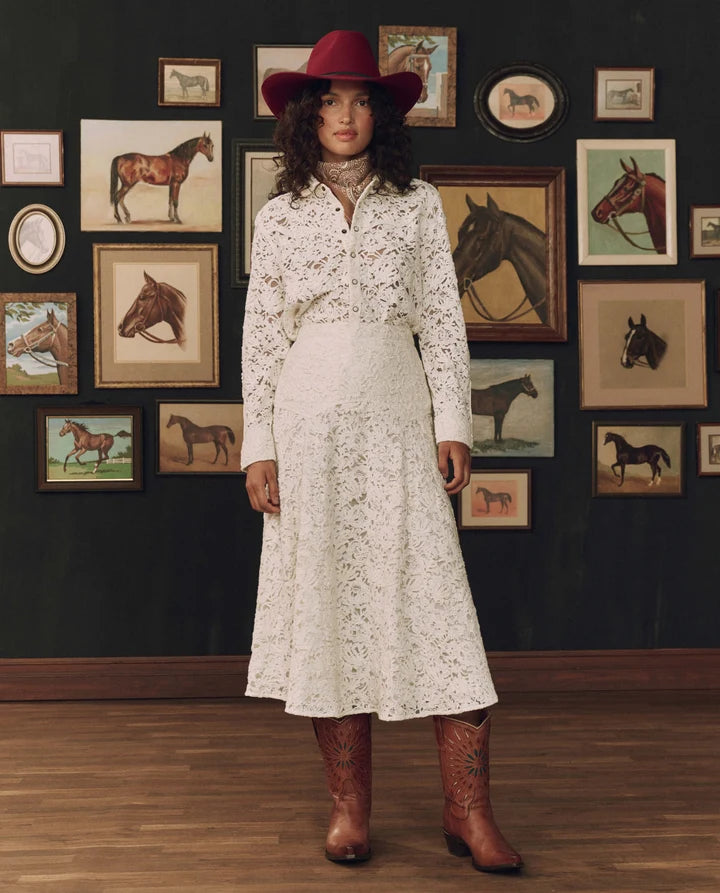 A woman stands in a room with a dark wall covered in framed horse illustrations, wearing a white long-sleeve floral set, a red hat, and brown cowboy boots.