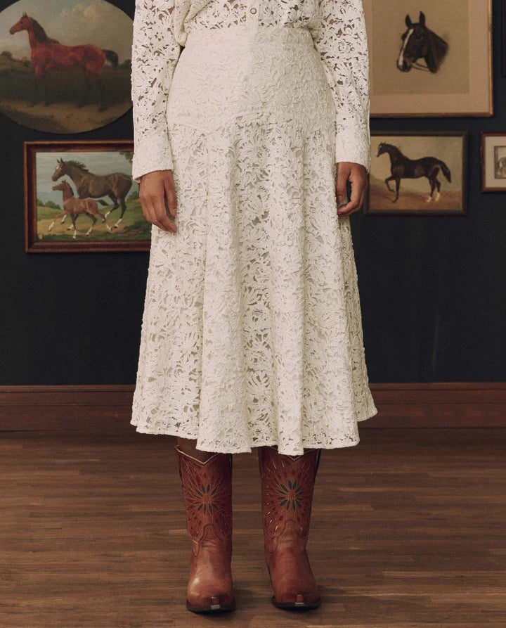 A person wearing a long-sleeve white lace midi skirt with a matching lace bodice, paired with brown western cowboy boots, standing in a room with horse-themed framed wall art.