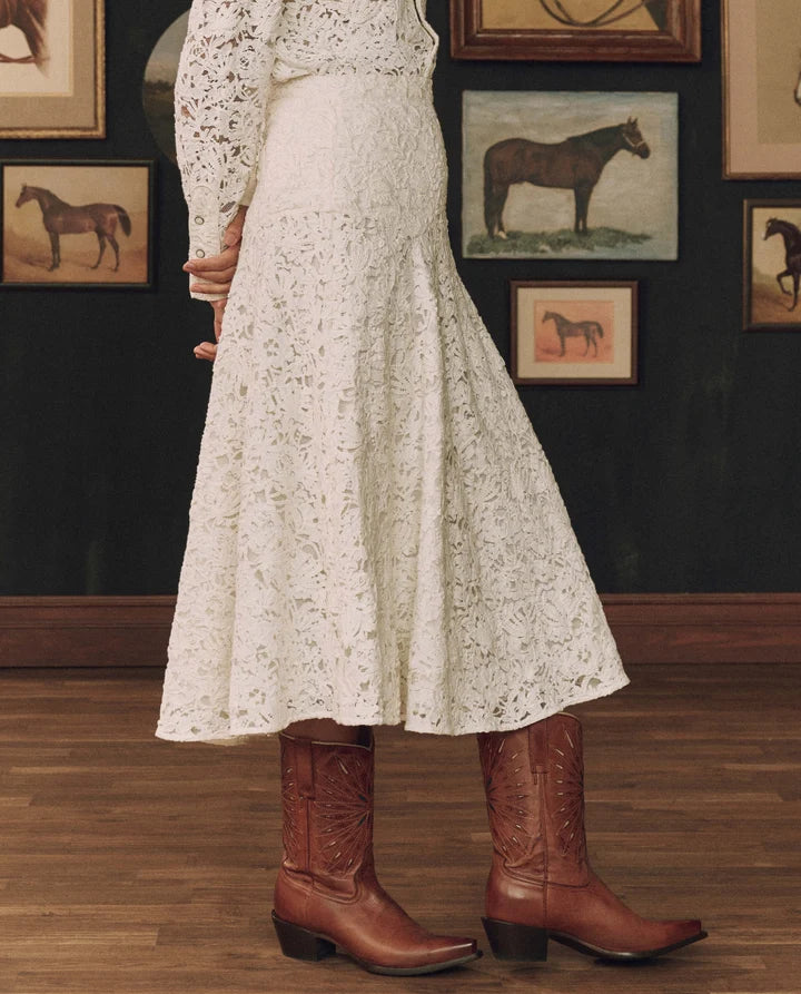 A person wearing a white floral lace midi skirt with a fitted bodice and long sleeves, paired with brown leather cowboy boots, standing in a room with framed horse paintings on a dark wall.