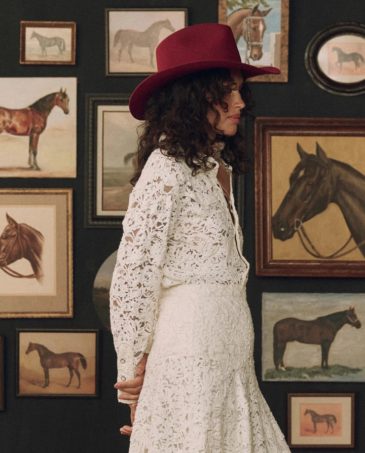 A woman in profile wearing a white lace long-sleeve dress and a dark red wide-brim hat, standing in front of a dark wall covered with framed horse paintings.