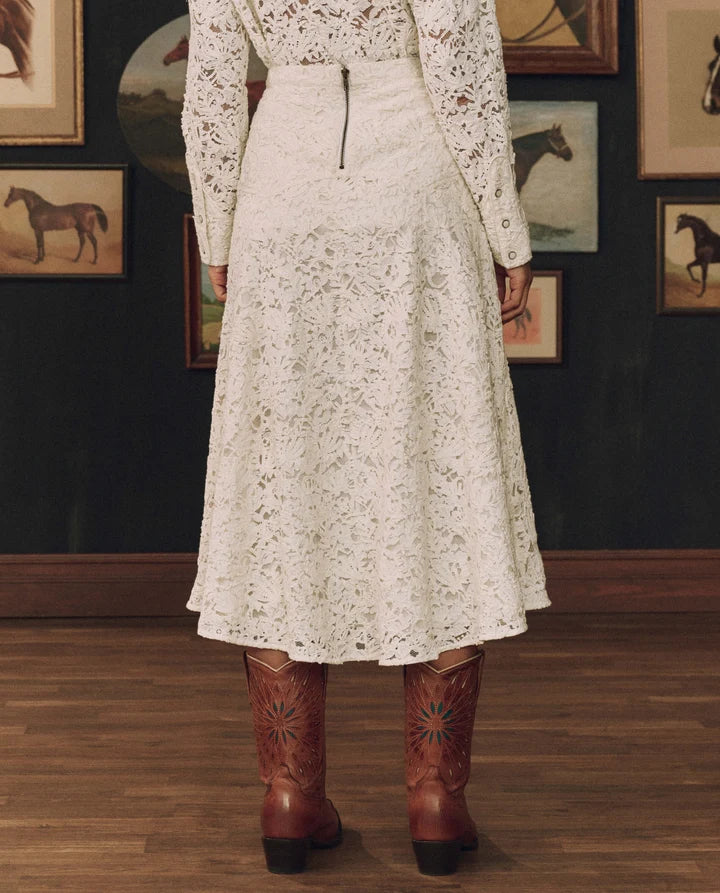 A person shown from the back wearing a cream lace long-sleeve midi dress with a zip closure, paired with brown western boots, standing in front of a dark wall with framed horse artwork.