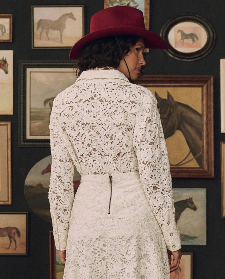 A woman shown from behind wearing a white lace long-sleeve dress and a dark red wide-brim hat, standing in front of a dark wall decorated with framed horse paintings.