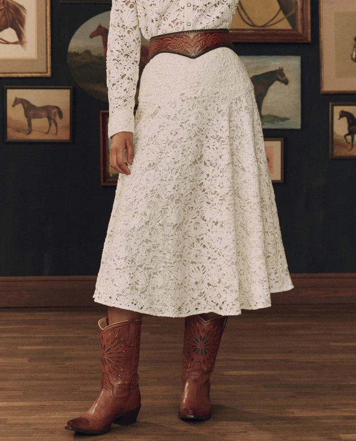 A person wearing a white floral lace midi skirt with a brown embossed leather belt, a matching white lace long-sleeve top, and brown leather cowboy boots, standing in a room with horse-themed framed wall art.