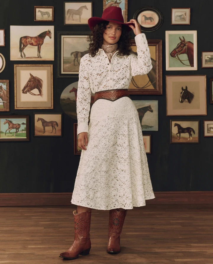 A woman stands in a room with a dark wall covered in framed horse illustrations, wearing a white floral long-sleeve shirt, a white lace midi skirt, a brown leather belt, a red hat, and brown cowboy boots.