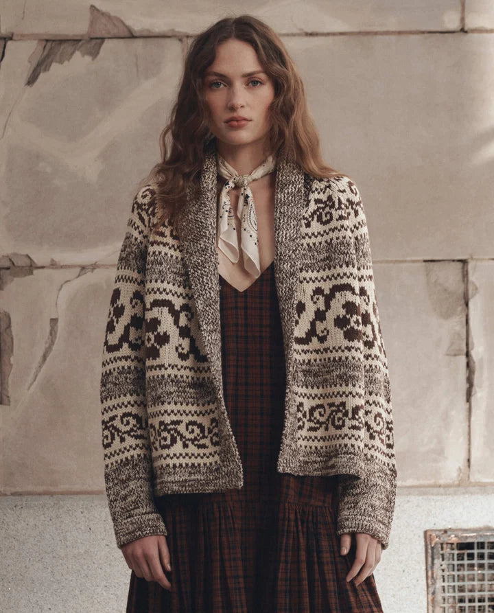 A woman stands against a textured stone wall wearing a cream and dark brown patterned knit cardigan over a dark red plaid dress, with a cream necktie.