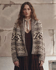 A woman stands against a textured stone wall wearing a cream and dark brown patterned knit cardigan over a dark red plaid dress, with a cream necktie.