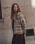 A woman wearing a cream and black patterned knit cardigan over a brown dress, standing against a textured concrete wall.