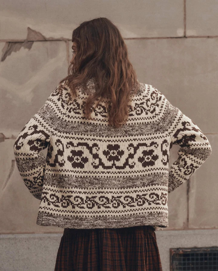 A woman viewed from behind wearing a cream and dark brown patterned knit sweater and a dark plaid skirt, standing against a textured concrete wall.