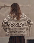 A woman viewed from behind wearing a cream and dark brown patterned knit sweater and a dark plaid skirt, standing against a textured concrete wall.
