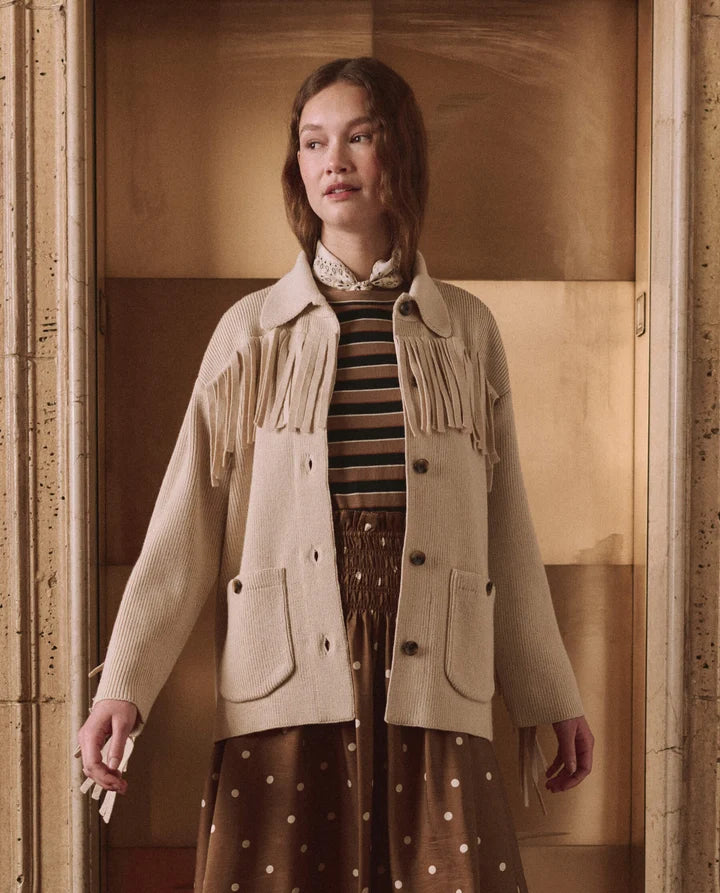 A model wearing a beige wool-blend fringe jacket over a striped top and a brown polka-dot skirt, standing in a warm-toned architectural setting.