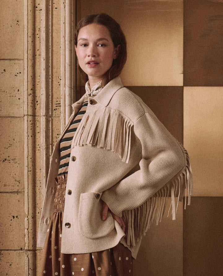 A woman models a cream-colored ribbed knit cardigan with long fringe detailing, styled over a striped shirt and a polka-dot skirt against a warm, checkered stone wall background.