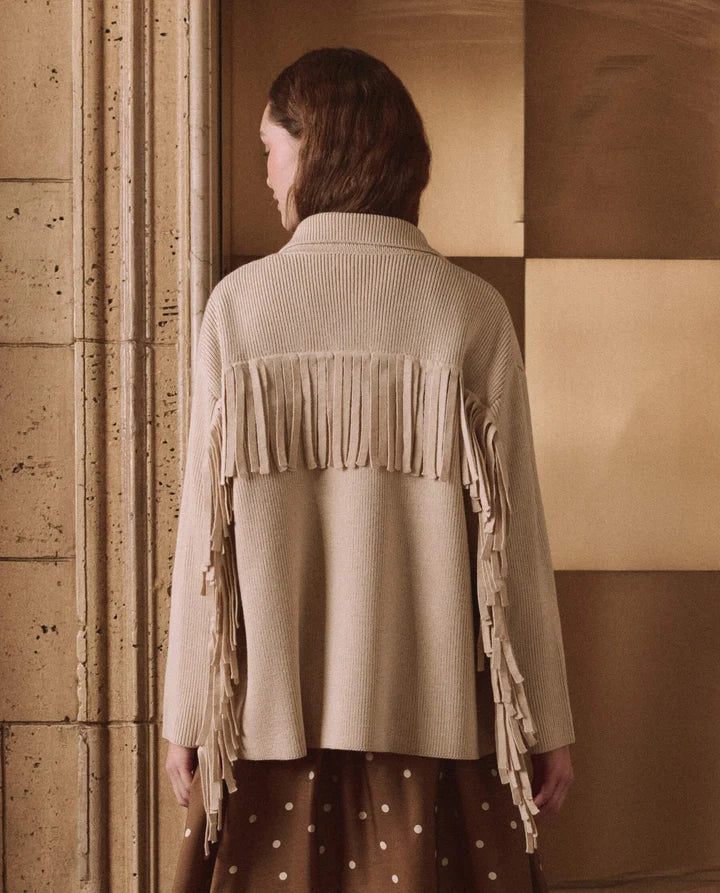 A woman shown from the back wearing a beige ribbed knit cardigan with long fringe across the upper back and hanging fringe down the sleeves, paired with a brown polka-dot skirt. She stands in front of a warm-toned paneled wall.