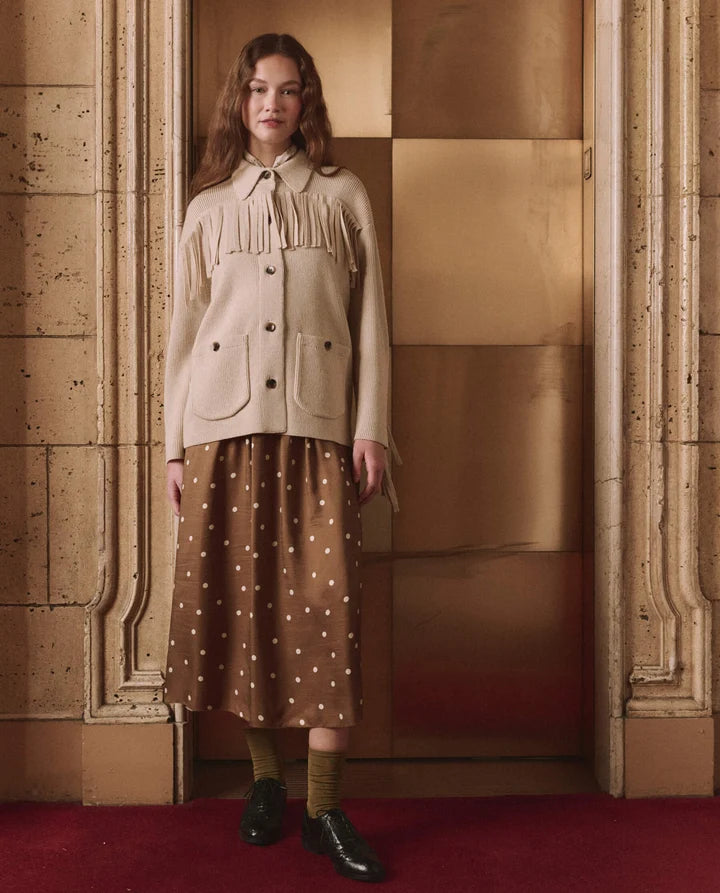A woman stands in a grand interior doorway wearing a cream fringe jacket with front pockets, a brown midi skirt with white polka dots, olive green socks, and black leather shoes.