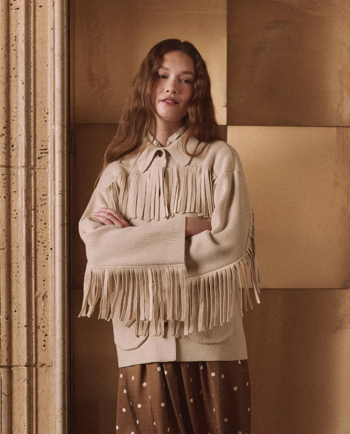 A woman wearing a beige fringed knit sweater over a brown polka dot top, standing against a warm beige paneled wall.