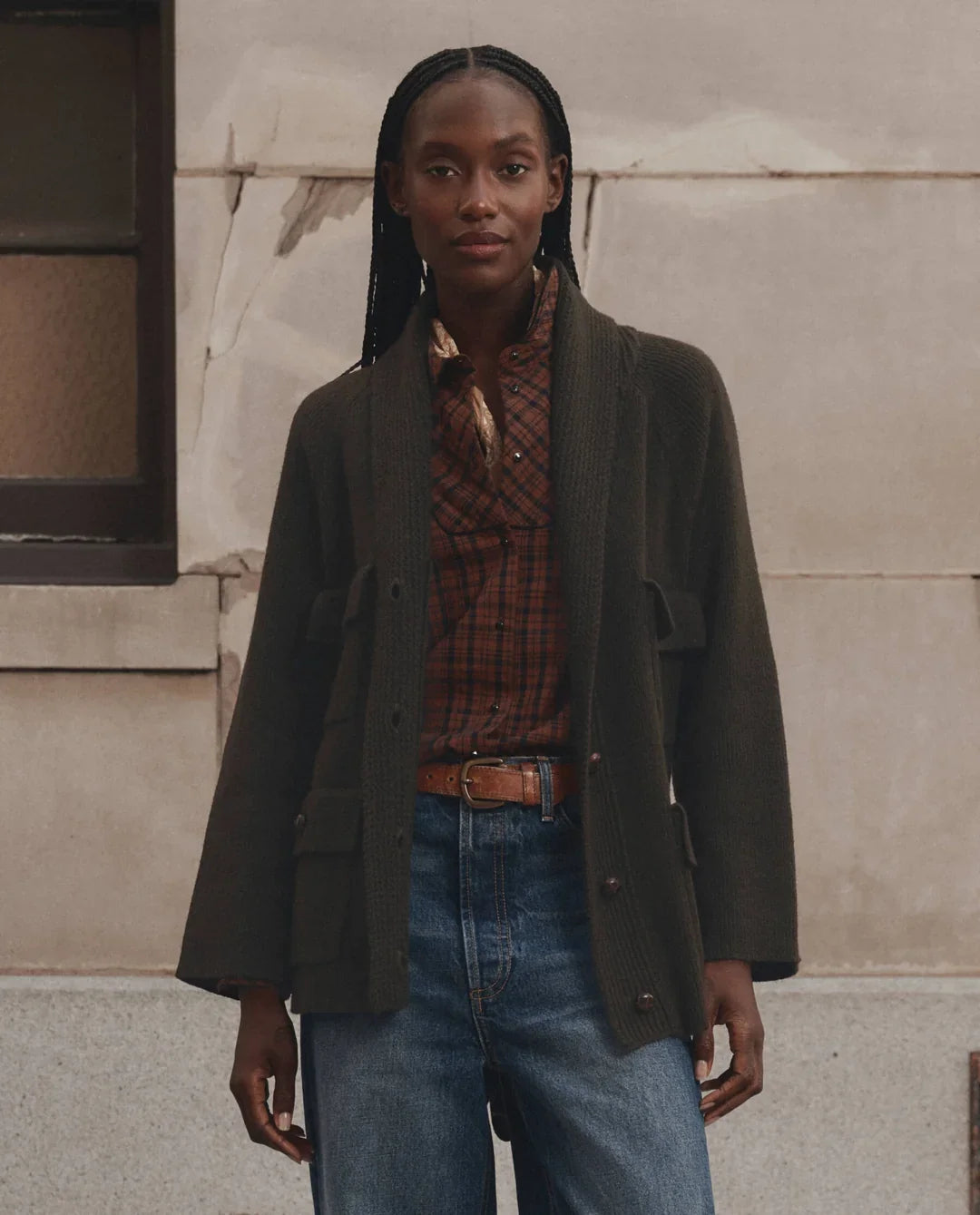 A model stands outdoors against a stone wall wearing a dark olive green open-front cardigan over a rust-red plaid shirt, paired with blue jeans and a brown belt.