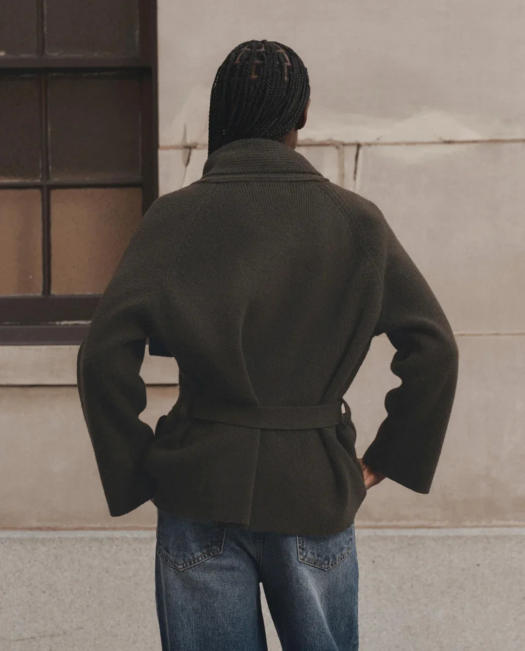 A person viewed from behind wearing an olive green belted coat and blue jeans, standing against a light beige stone wall.