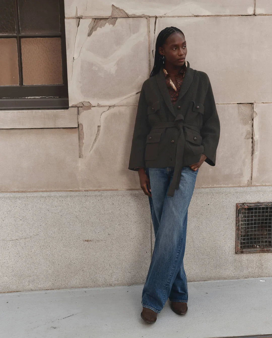 A woman stands against a textured beige stone wall wearing a dark olive green belted jacket, blue wide-leg jeans, and brown shoes.
