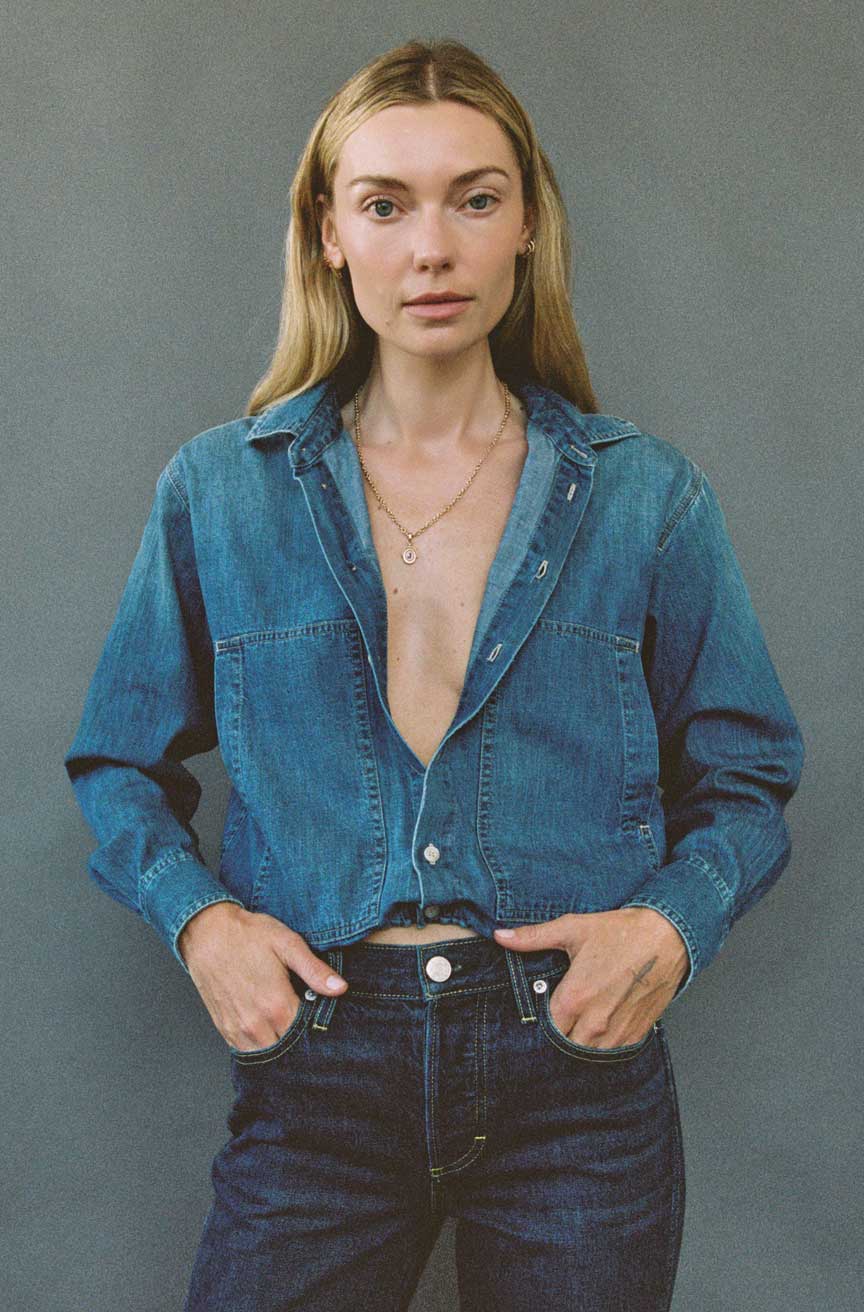 A woman modeling a cropped blue denim shirt-jacket with a deep V-neck over dark blue jeans, standing against a solid gray studio background.
