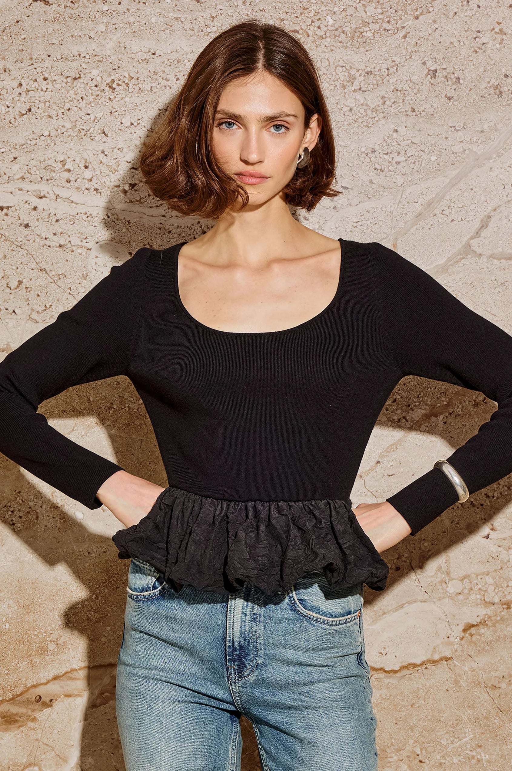 Woman wearing a black top with ruffled hem and blue jeans against a stone wall front view