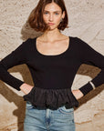 Woman wearing a black top with ruffled hem and blue jeans against a stone wall front view