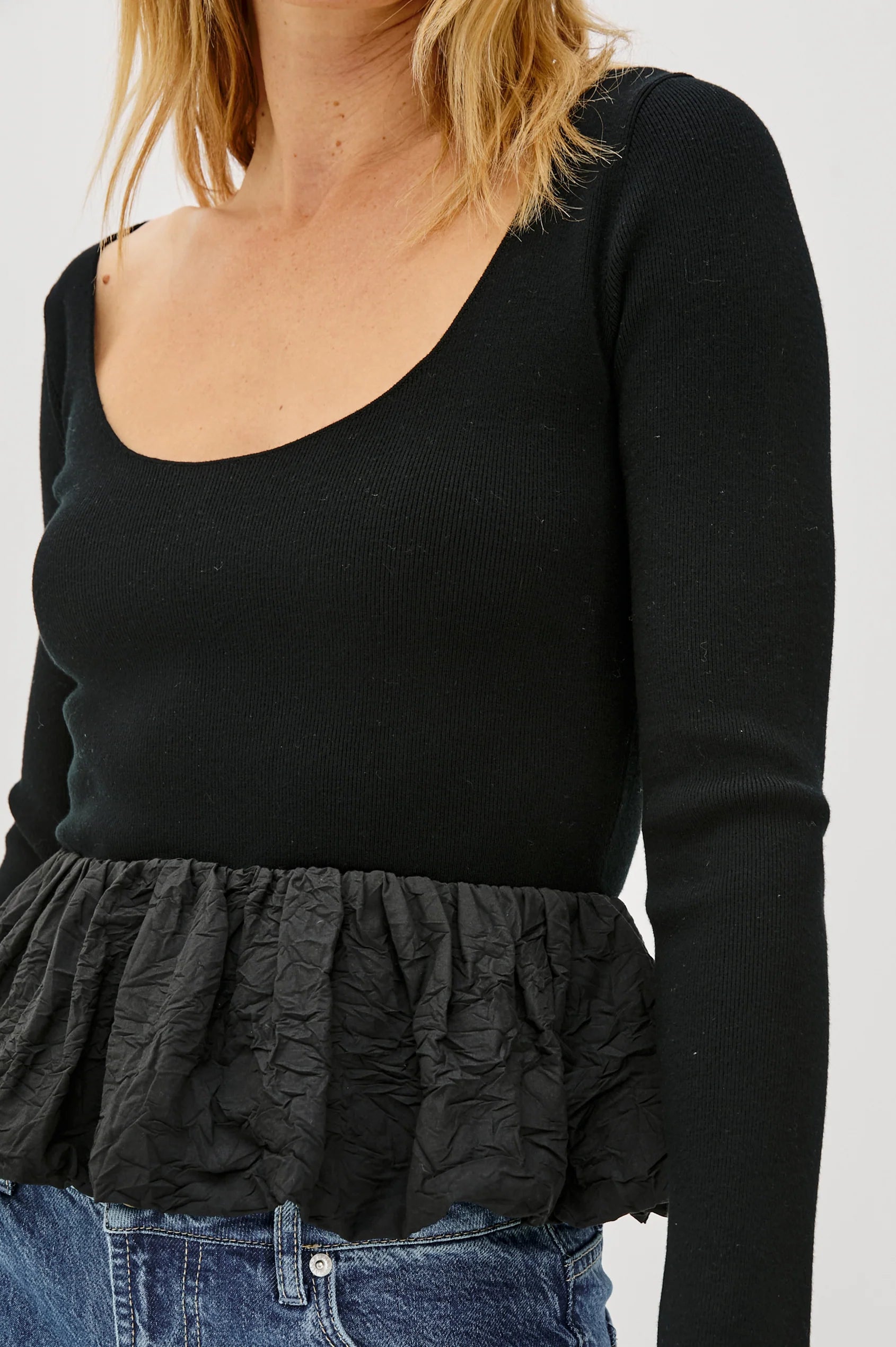 Black long-sleeve top with a ruffled bottom worn by a person on a white background detail view