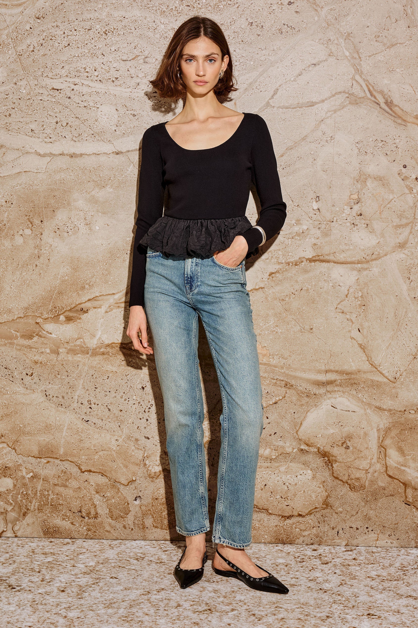 Person wearing a black long-sleeve top with a peplum waist and blue jeans on a stone background front view