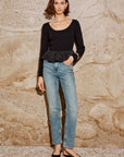 Person wearing a black long-sleeve top with a peplum waist and blue jeans on a stone background front view