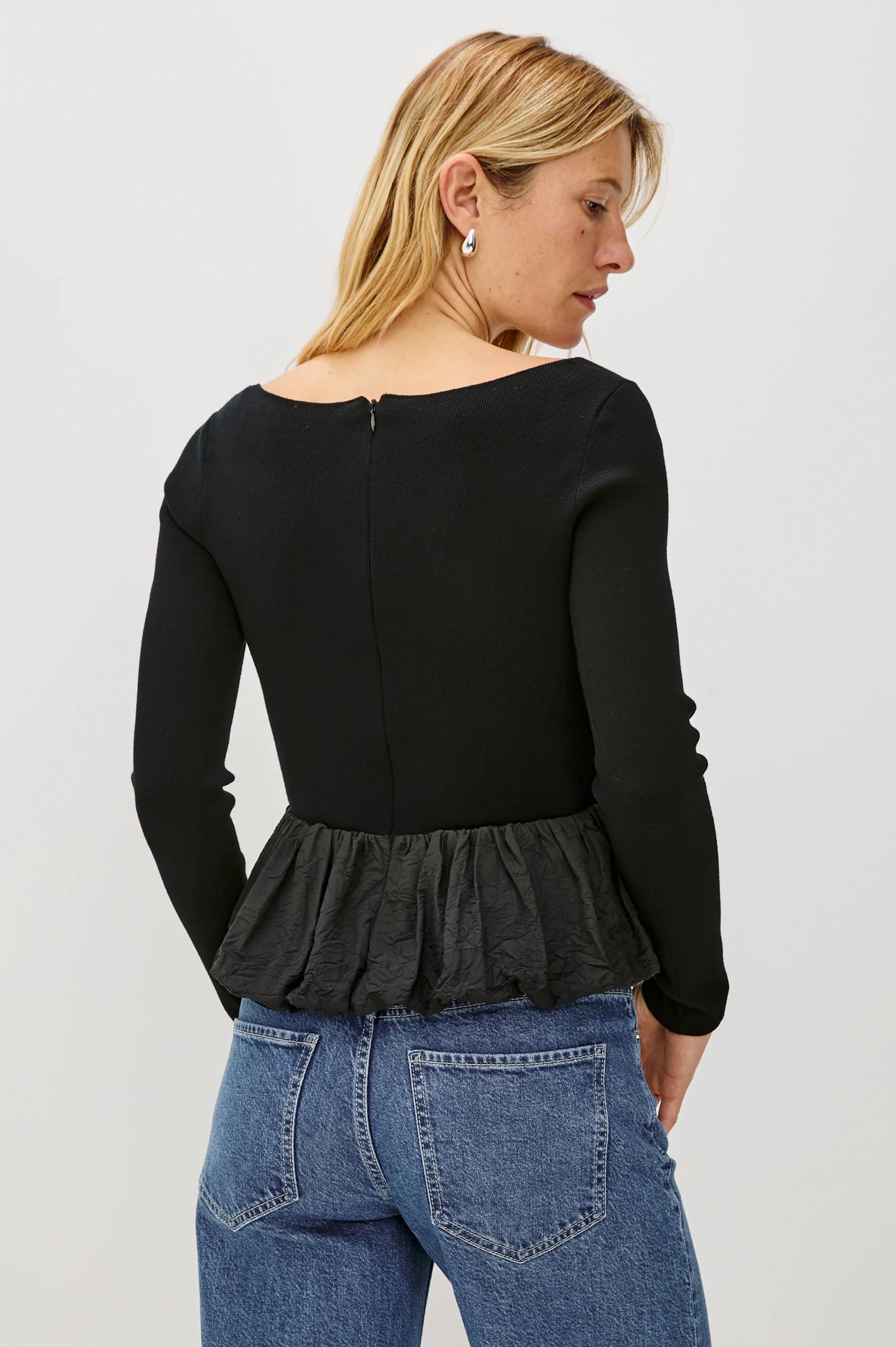 Person wearing a black long-sleeve top with a peplum waist and blue jeans on a white background back view