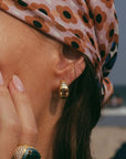 A close-up lifestyle shot of a woman wearing a gold huggie hoop earring and a matching gold ring with a green stone, styled with a pink and brown patterned headscarf on a beach background.