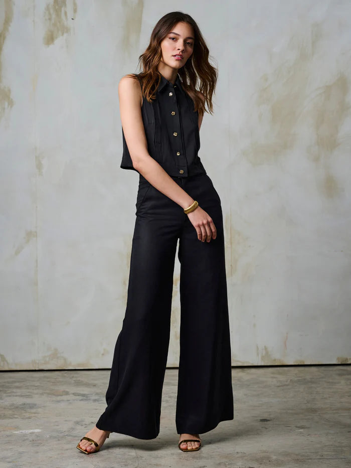 Woman wearing a black sleeveless top and wide-leg pants against a textured wall.