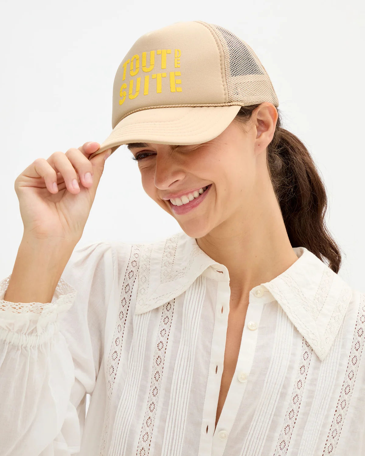 A woman wearing a beige trucker-style cap with yellow embroidered text reading 'TOUT DE SUITE' and a white lace blouse, photographed against a clean white background.